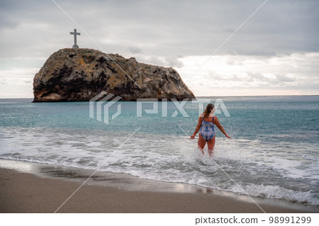 A plump woman in a bathing suit enters the water during the surf. Alone on the beach, Gray sky in the clouds, swimming in winter. A plump woman in a bathing suit enters the water during the surf. Alone on the beach, Gray sky in the clouds, swimming in winter. 98991299