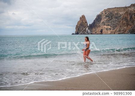 A plump woman in a bathing suit enters the water during the surf. Alone on the beach, Gray sky in the clouds, swimming in winter. A plump woman in a bathing suit enters the water during the surf. Alone on the beach, Gray sky in the clouds, swimming in winter. 98991300