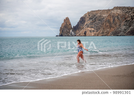 A plump woman in a bathing suit enters the water during the surf. Alone on the beach, Gray sky in the clouds, swimming in winter. A plump woman in a bathing suit enters the water during the surf. Alone on the beach, Gray sky in the clouds, swimming in winter. 98991301