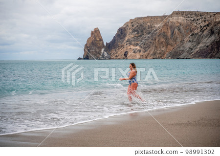 A plump woman in a bathing suit enters the water during the surf. Alone on the beach, Gray sky in the clouds, swimming in winter. A plump woman in a bathing suit enters the water during the surf. Alone on the beach, Gray sky in the clouds, swimming in winter. 98991302
