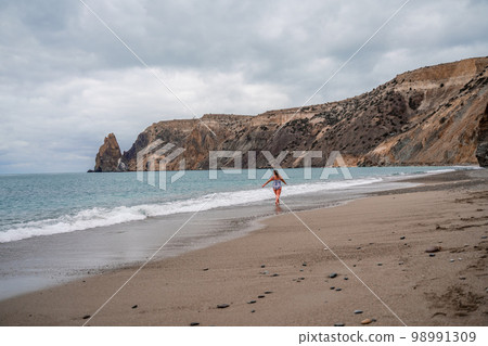 A plump woman in a bathing suit enters the water during the surf. Alone on the beach, Gray sky in the clouds, swimming in winter. A plump woman in a bathing suit enters the water during the surf. Alone on the beach, Gray sky in the clouds, swimming in winter. 98991309