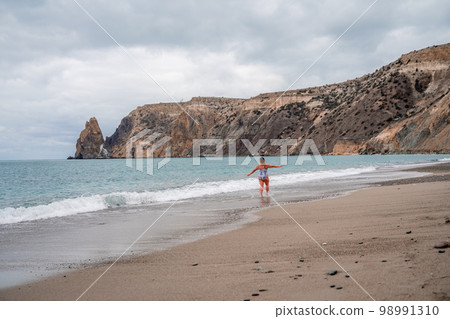 A plump woman in a bathing suit enters the water during the surf. Alone on the beach, Gray sky in the clouds, swimming in winter. 98991310