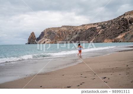 A plump woman in a bathing suit enters the water during the surf. Alone on the beach, Gray sky in the clouds, swimming in winter. 98991311