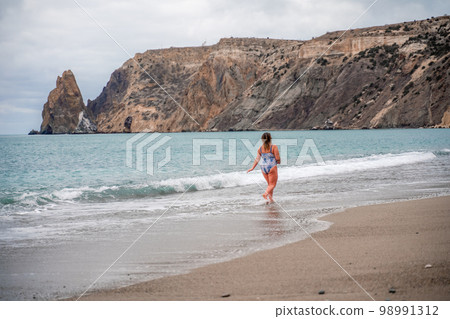 A plump woman in a bathing suit enters the water during the surf. Alone on the beach, Gray sky in the clouds, swimming in winter. 98991312