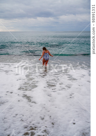 A plump woman in a bathing suit enters the water during the surf. Alone on the beach, Gray sky in the clouds, swimming in winter. 98991331