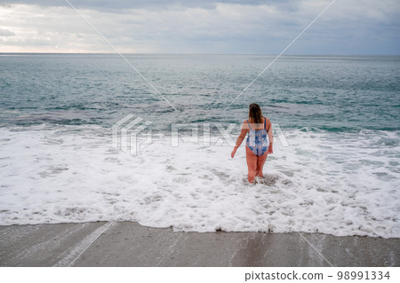 A plump woman in a bathing suit enters the water during the surf. Alone on the beach, Gray sky in the clouds, swimming in winter. 98991334