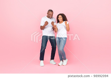 Cheerful black couple gambling together online using smartphone, rasising hands up and smiling, pink studio background 98992094