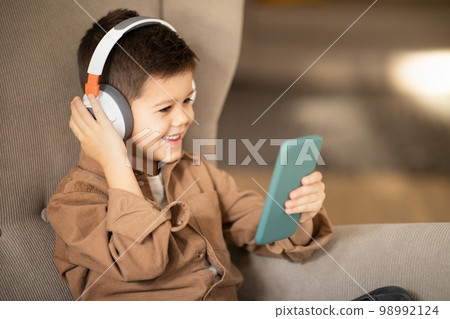 Happy european little boy in casual and wireless headphones watch video on smartphone, sits in armchair Happy european little boy in casual and wireless headphones watch video on smartphone, sits in armchair 98992124