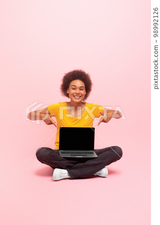 Cheerful young african american curly woman sit on floor, point fingers at laptop with blank screen Cheerful young african american curly woman sit on floor, point fingers at laptop with blank screen 98992126