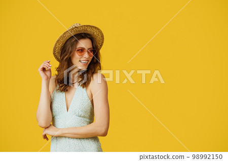 Summer travels. Young woman in dress, straw hat and colorful sunglasses looking aside at copy space, yellow background Summer travels. Young woman in dress, straw hat and colorful sunglasses looking aside at copy space, yellow background 98992150