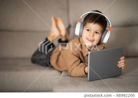Smiling caucasian little kid in headphones enjoys free time, watching video, playing game on tablet Smiling caucasian little kid in headphones enjoys free time, watching video, playing game on tablet 98992200