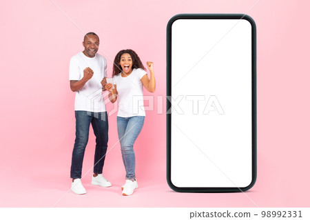 Excited black couple near big cellphone empty screen gesturing yes and exclaiming on pink background, full length Excited black couple near big cellphone empty screen gesturing yes and exclaiming on pink background, full length 98992331