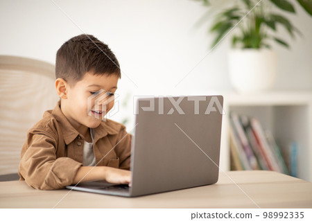 Glad surprised cute caucasian little child pupil sits at table, typing on computer, enjoy lesson in room 98992335