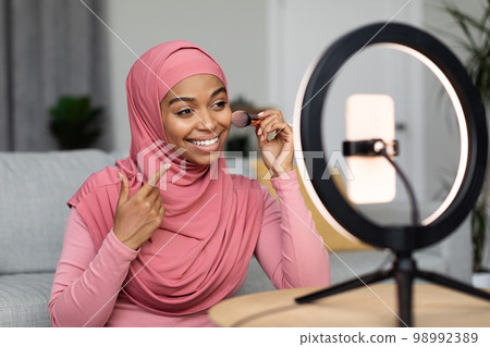 Cheerful black muslim woman recording cosmetics product review, applying powder, filming video for her vlog using phone 98992389