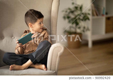 Surprised smiling cute caucasian little kid with smartphone sits in armchair and looks at empty space 98992420