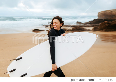 Happy surfer girl walking with board on the sandy beach, looking away and smiling, free space 98992450