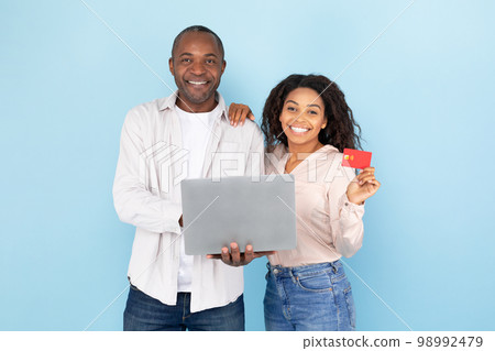 Happy black middle aged man and young woman showing credit card and laptop, standing over blue studio background 98992479