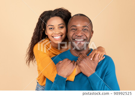 Portrait of happy young black woman hugging her middle aged husband from behind over peach studio background 98992525