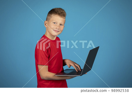 Smiling Preteen Boy Using Laptop Computer While Standing Over Blue Studio Background Smiling Preteen Boy Using Laptop Computer While Standing Over Blue Studio Background 98992538