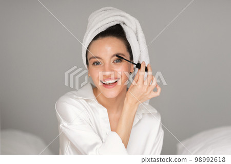 Cheerful young european lady in towel applies mascara on eyelashes, enjoys daily procedures 98992618