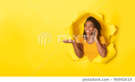Excited black lady talking on cellphone and looking at free space, posing in torn yellow paper background, panorama 98992619