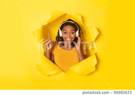 Happy black lady in wireless headphones listening to music online and looking at camera through hole in torn paper 98992623