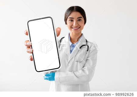 Young Indian Female Doctor In Uniform Showing Big Smartphone With Blank Screen Young Indian Female Doctor In Uniform Showing Big Smartphone With Blank Screen 98992626