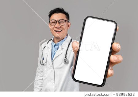 Asian male doctor showing smartphone with big blank screen at camera Asian male doctor showing smartphone with big blank screen at camera 98992628