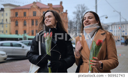 Two joyful friends in a jacket and coat are walking around the city with tulips. Two joyful friends in a jacket and coat are walking around the city with tulips. 98992743