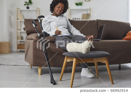 Attractive smiling african american woman works at home in the living room with laptop and makes notes in notebook, sitting on the sofa with broken bandaged leg lying on stool. Attractive smiling african american woman works at home in the living room with laptop and makes notes in notebook, sitting on the sofa with broken bandaged leg lying on stool. 98992914