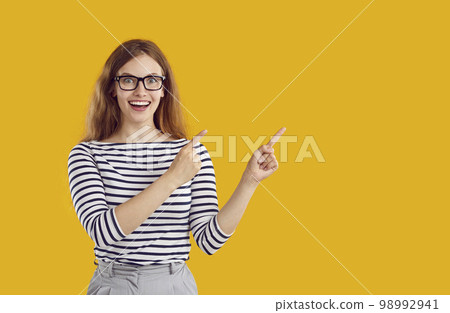 Young excited woman in glasses with wavy hair is pointing on copy space on yellow background. Young excited woman in glasses with wavy hair is pointing on copy space on yellow background. 98992941