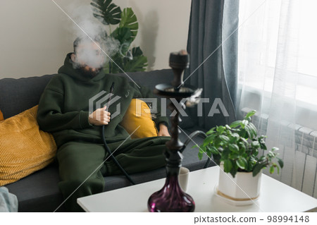 Bearded man is smoking hookah at home and blowing cloud of smoke - chill time and resting concept Bearded man is smoking hookah at home and blowing cloud of smoke - chill time and resting concept 98994148