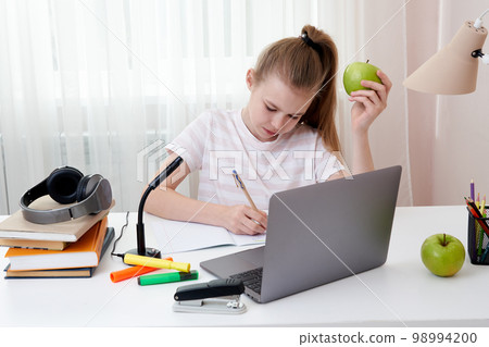 Teenage girl holding green apple studying at home with laptop Teenage girl holding green apple studying at home with laptop 98994200