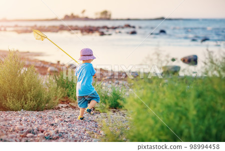 Child going to the water with net in his hand along pebble seaside. 98994458