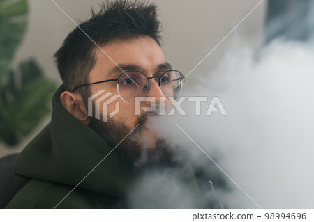 Portrait bearded man is smoking hookah at home and blowing cloud of smoke - chill time and resting concept Portrait bearded man is smoking hookah at home and blowing cloud of smoke - chill time and resting concept 98994696