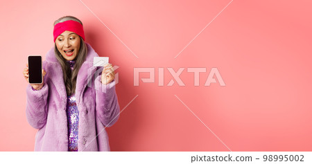 Special promotion. Excited asian senior woman showing blank smartphone screen and plastic credit card, checking out online offer, pink background 98995002