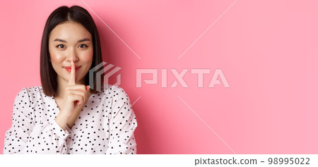 Close-up of mysterious asian woman hiding a secret, hushing and telling to keep quiet, standing over pink background 98995022