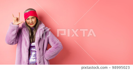 Funny and cool asian senior woman showing rock n roll gesture, looking sassy, standing over pink background 98995023