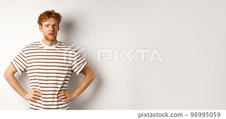 Confused and offended man with ginger curly hair staring at camera puzzled, standing questioned against white background Confused and offended man with ginger curly hair staring at camera puzzled, standing questioned against white background 98995059