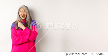 Portrait of impressed asian lady checking out promo offer, pointing at upper left corner and staring at camera amazed, standing in trendy pink sweater over white background 98995063