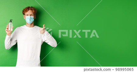 Covid-19, virus and social distancing concept. Smiling young man with red hair, wearing face mask for protection from corona, showing thumb-up and hand sanitizer, green background 98995065