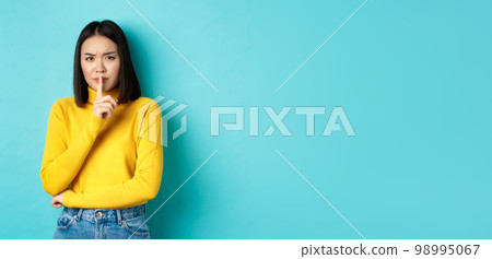 Disappointed asian woman telling to be quiet, scolding loud person with hush gesture, shushing at camera and frowning upset, standing over blue background Disappointed asian woman telling to be quiet, scolding loud person with hush gesture, shushing at camera and frowning upset, standing over blue background 98995067