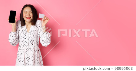 Joyful korean woman showing smartphone screen and credit card, paying for internet order, demonstrating online shopping app, standing over pink background 98995068