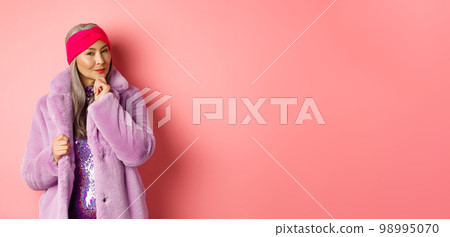 Fashion and shopping concept. Stylish old asian lady in purple fake-fur coat looking intrigued, interested with promotion, smiling and thinking, pink background Fashion and shopping concept. Stylish old asian lady in purple fake-fur coat looking intrigued, interested with promotion, smiling and thinking, pink background 98995070