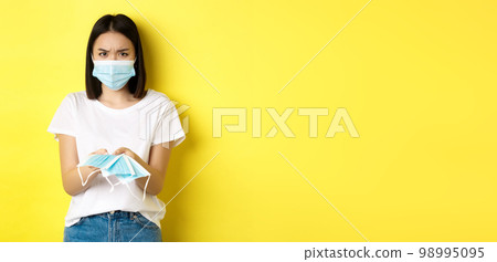 Coronavirus, quarantine and medicine concept. Angry asian girl giving you medical mask indoors, frowning upset, standing over yellow background 98995095