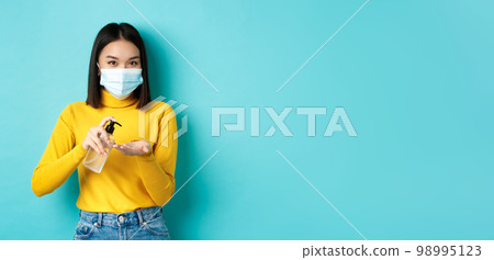 Covid-19, social distancing and pandemic concept. Smiling asian woman in medical mask using preventive measures from coronavirus, using hand sanitizer, standing over blue background 98995123