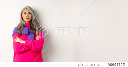 Serious and strict asian grandmother looking angry at camera, cross arms on chest, standing over white background disappointed 98995125