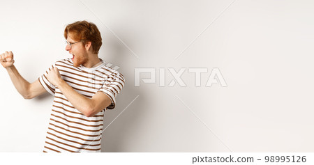 Profile of funny young man with red hair and beard, raising fists for fight, shadow boxing and shouting, standing over white background 98995126