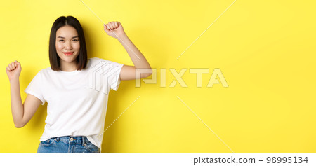 Carefree asian girl dancing and having fun, posing in white t-shirt against yellow background Carefree asian girl dancing and having fun, posing in white t-shirt against yellow background 98995134