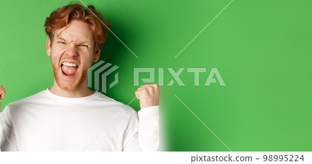 Close-up of lucky young man winner, celebrating victory, shouting yes with joy and making fist pumps, triumphing on green background 98995224